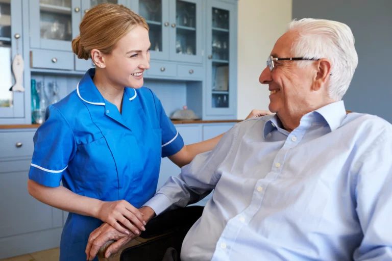 A nurse in a blue uniform smiling and talking with an elderly person in a blue shirt. They are indoors with blue kitchen cabinets in the background
