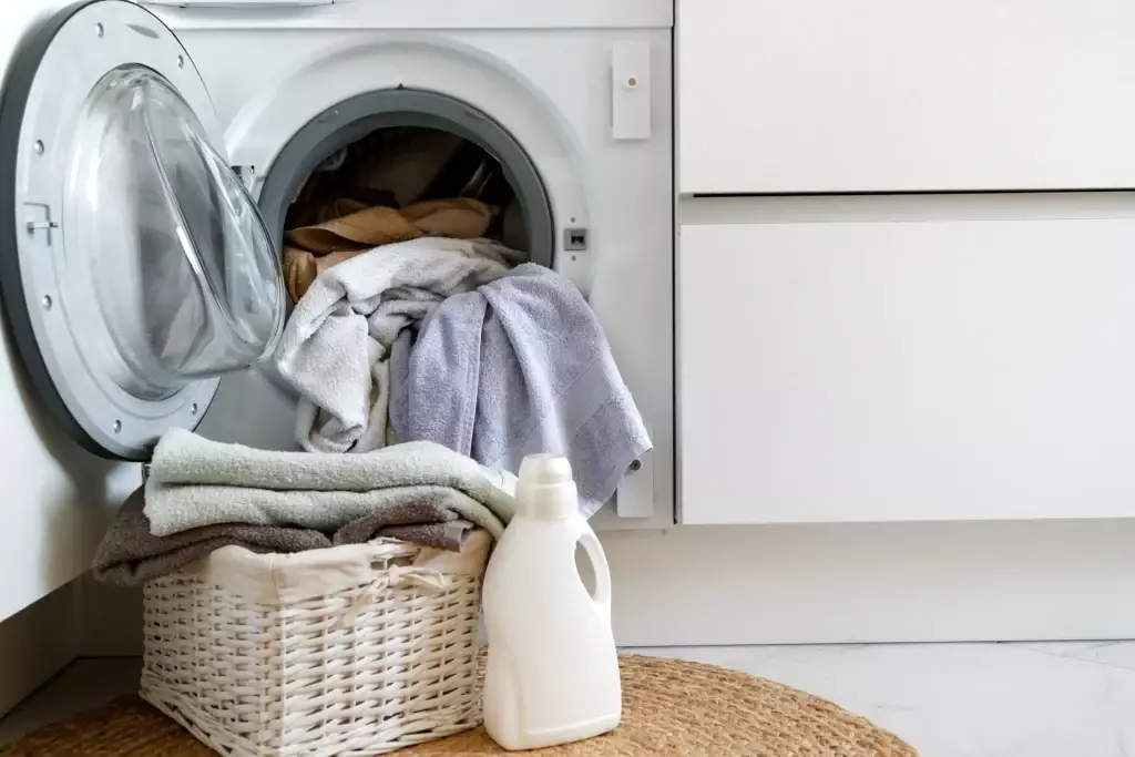 A half-loaded washing machine with an open door. There are towels draped in the machine while a basket of folded towels and a bottle of laundry detergent sits in front