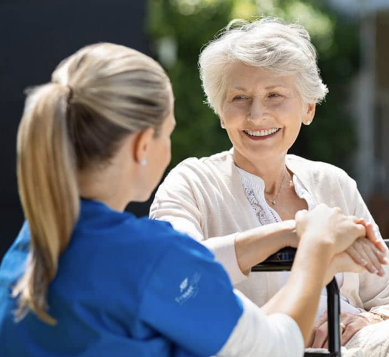 A Fraser Elite care worker in a blue uniform providing support to a smiling elderly woman sitting down outdoors
