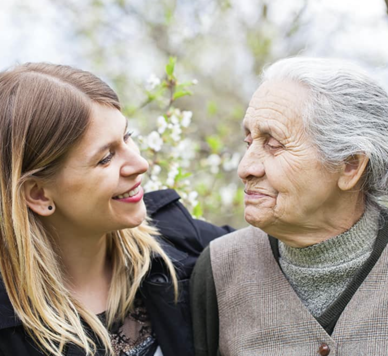 A close-up of two people smiling at each other outdoors - one younger person with long hair and an elderly person, with leaves and flowers in the background