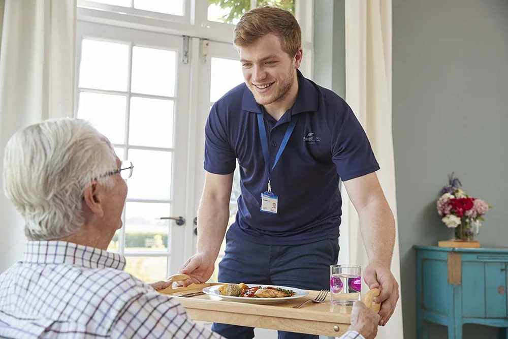 A smiling care worker for Fraser Elite dressed in navy blue serving a meal tray of food to an elderly person