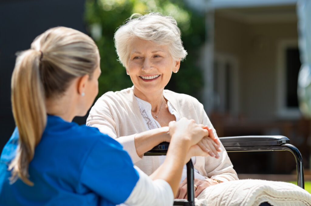 A care support worker in a blue uniform assisting and holding the hand of a smiling elderly person who is sitting in a wheelchair outdoors