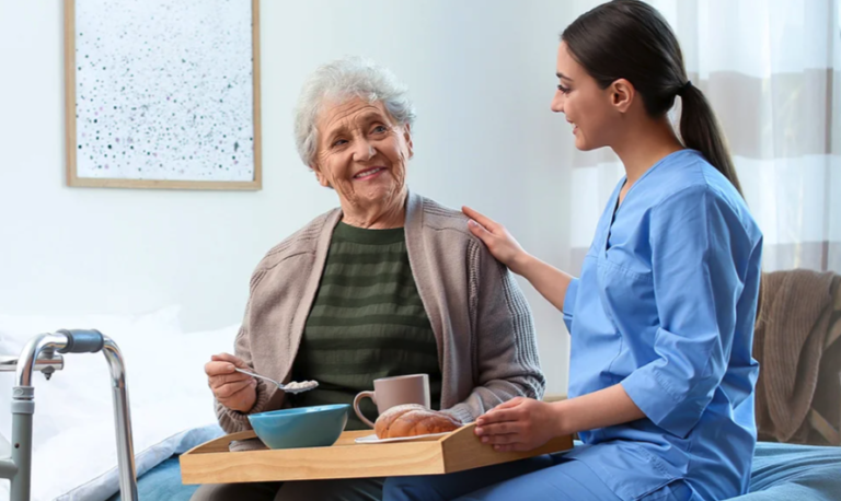 A care worker in a blue uniform smiling and touching the shoulder of an elderly person next to her. The elderly person has a tray of food in her lap