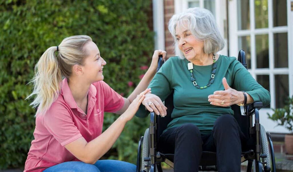 A Fraser Elite care worker kneeling next to an elderly person in a wheelchair, talking and smiling together outdoors