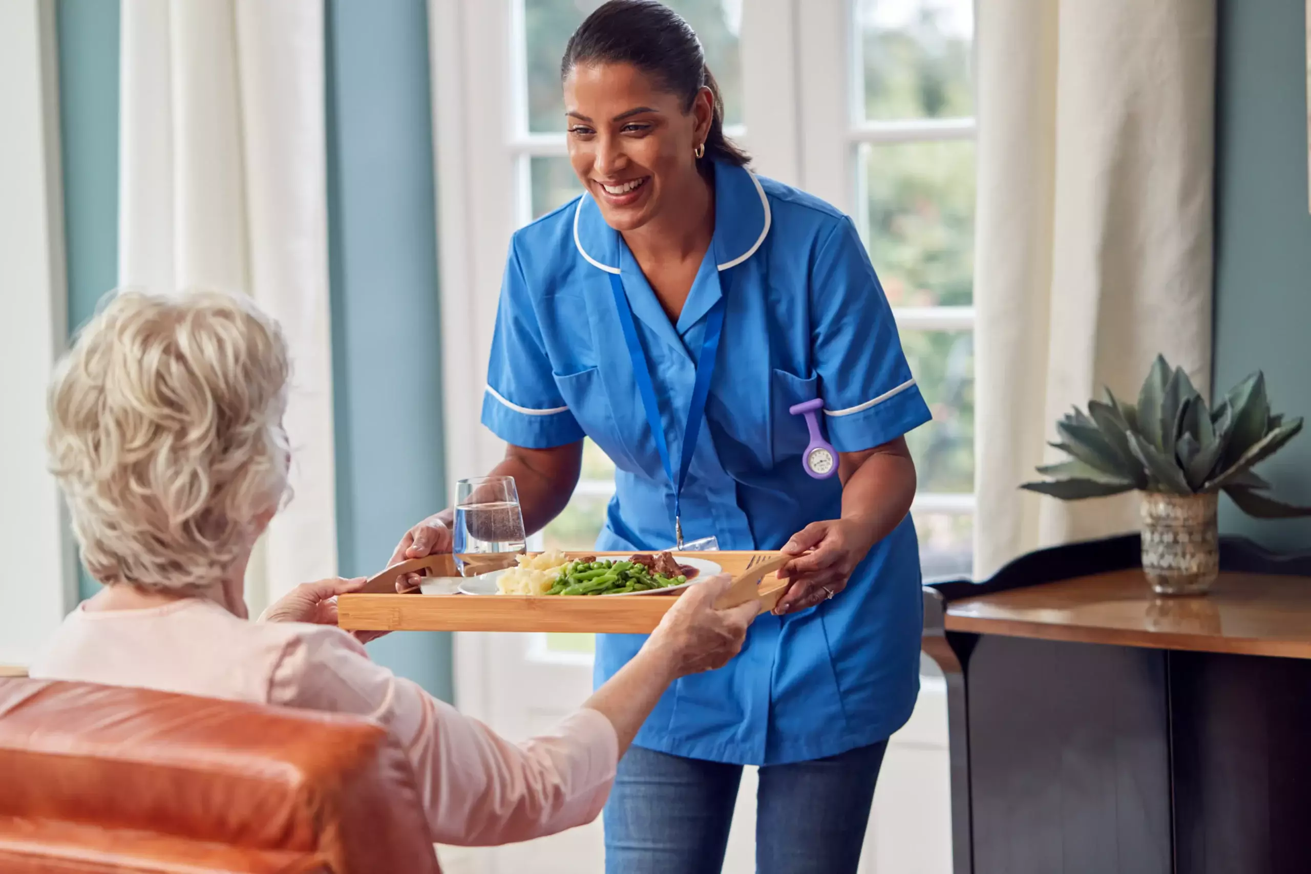 A care worker in a light blue uniform and jeans smiling and serving a tray of food to an elderly person at home