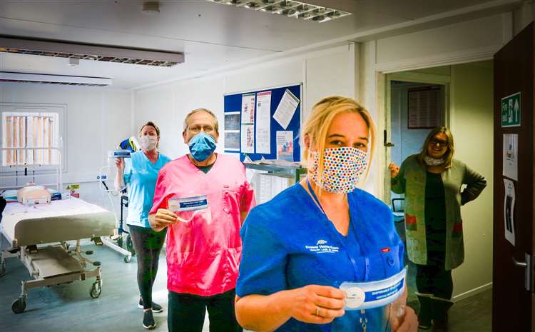Three care nurses standing in single file wearing face masks and posing for the camera next to a smiling person in the doorway. A hospital bed sits in the background
